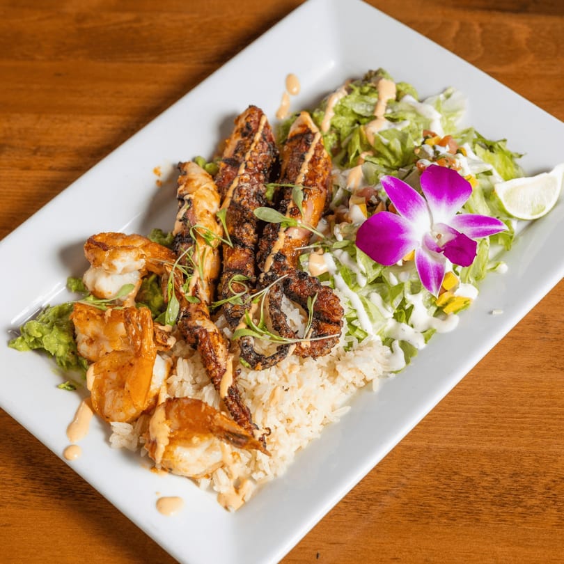 Grilled shrimp and octopus with rice, guacamole, salad, and a purple orchid on a white plate.