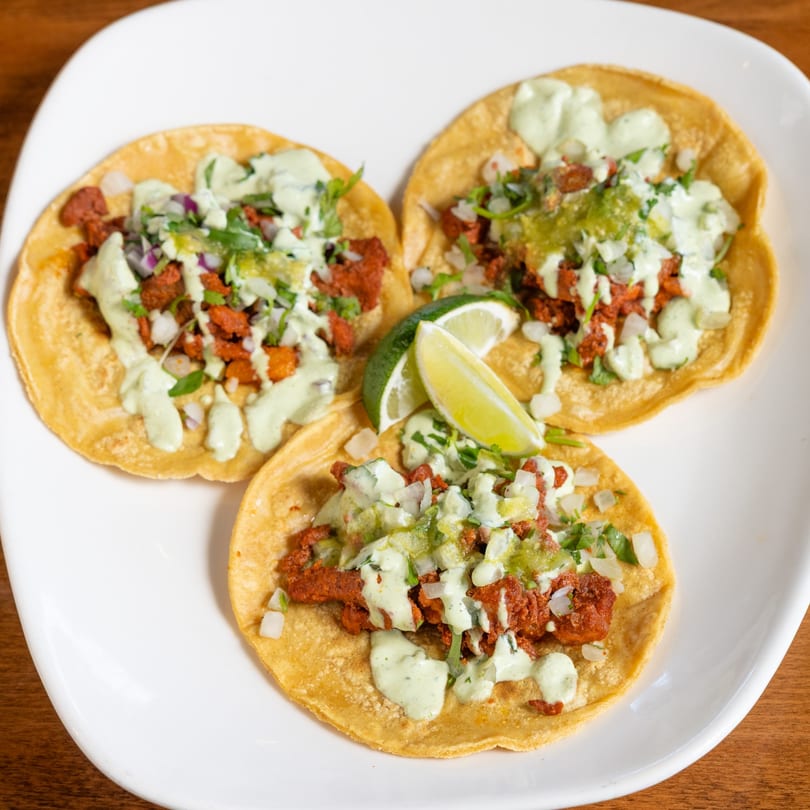 Three California Tacos with red meat, green sauce, onions, cilantro, and lime wedges on a white plate.