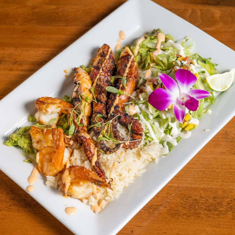 Grilled shrimp and octopus with rice, guacamole, salad, and a purple orchid on a white plate.