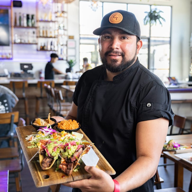 A chef in a California Tacos hat holds a wooden board with tacos, rice, and dip.