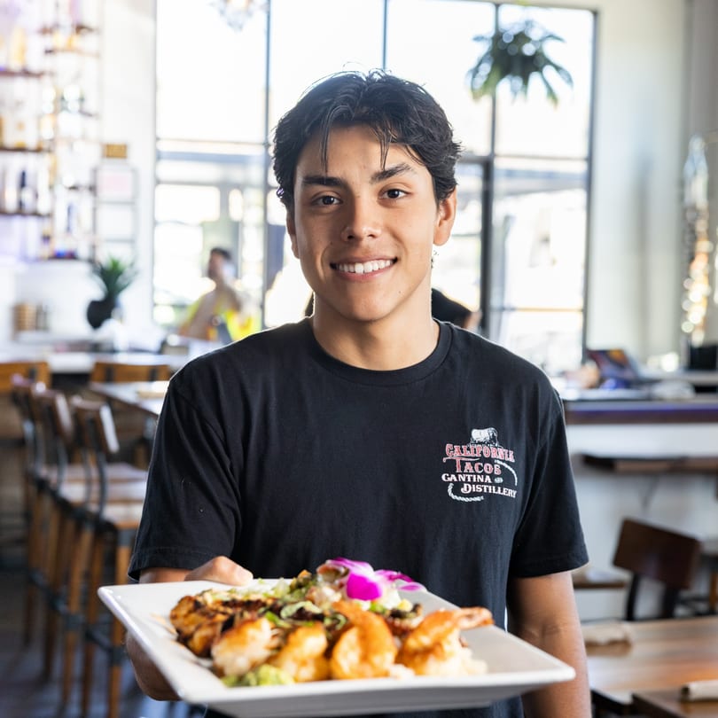Smiling California Tacos Cantina and Distillery server holding a plate of shrimp and steak.