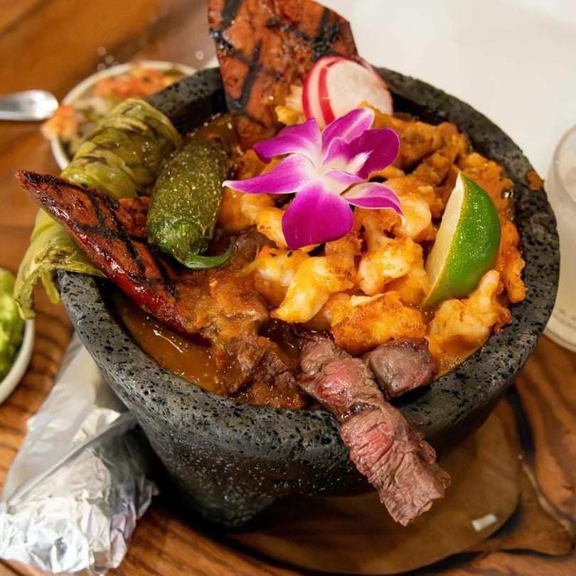 Molcajete with grilled meats, shrimp, peppers, and a purple orchid garnish at California Tacos.