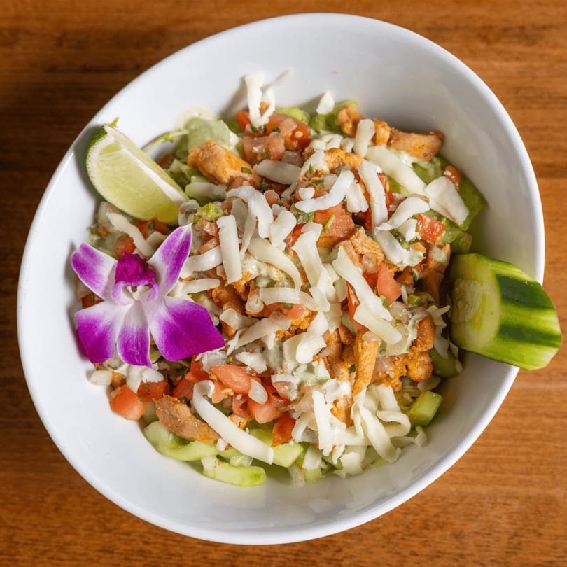 Mexican salad with chicken, cheese, tomatoes, and a purple orchid in a white bowl.