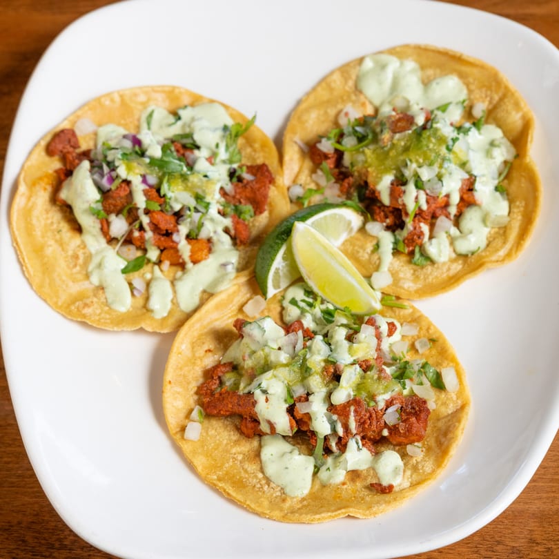 Three California Tacos al pastor with green sauce, onions, cilantro, and lime wedges on a white plate.