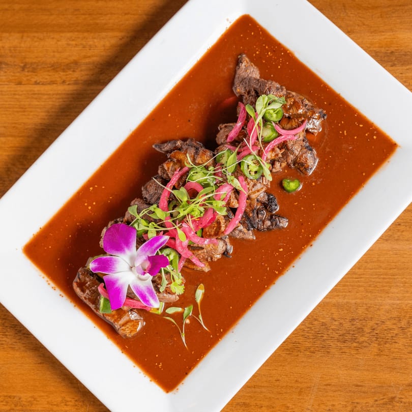 Grilled steak in a rich red sauce, topped with pickled red onions, jalapeños, and a purple orchid.